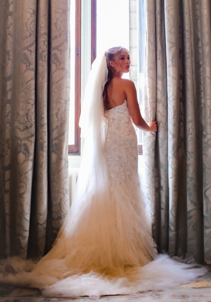 Bride in wedding dress, standing at window.