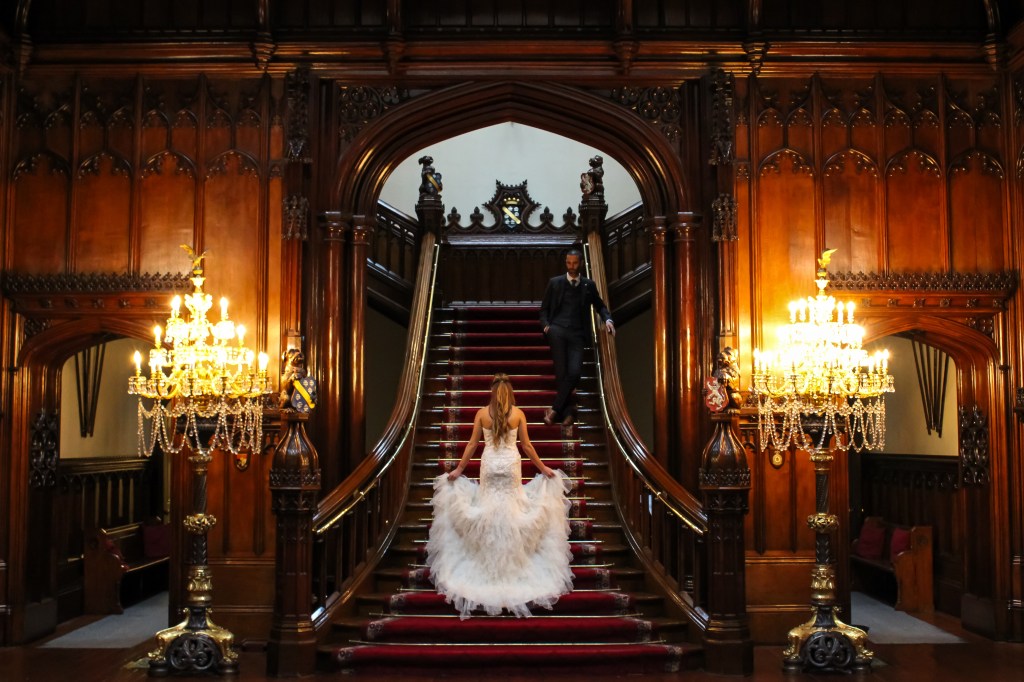 Bride in wedding dress, standing on staircase, looking up to the waiting groom.