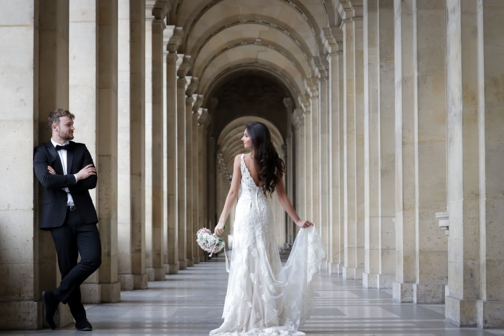 Groom leaning against wall while bride in wedding dress looks on underneath arches.